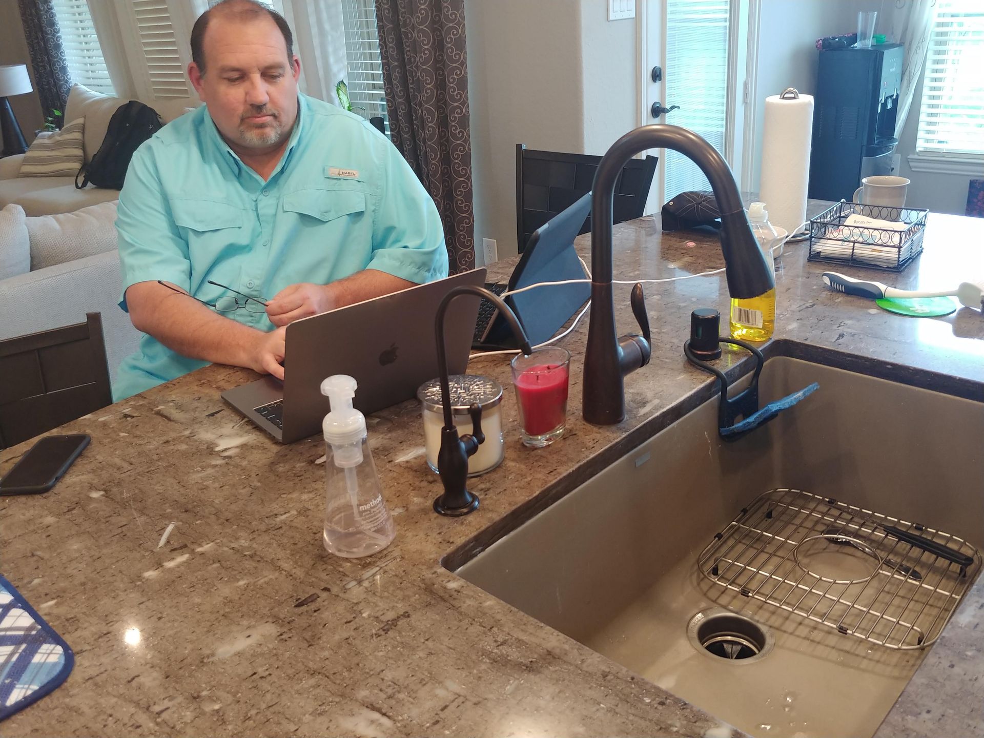 Man works on a laptop at a kitchen counter, near a sink. He has a serious expression, and other items are nearby.