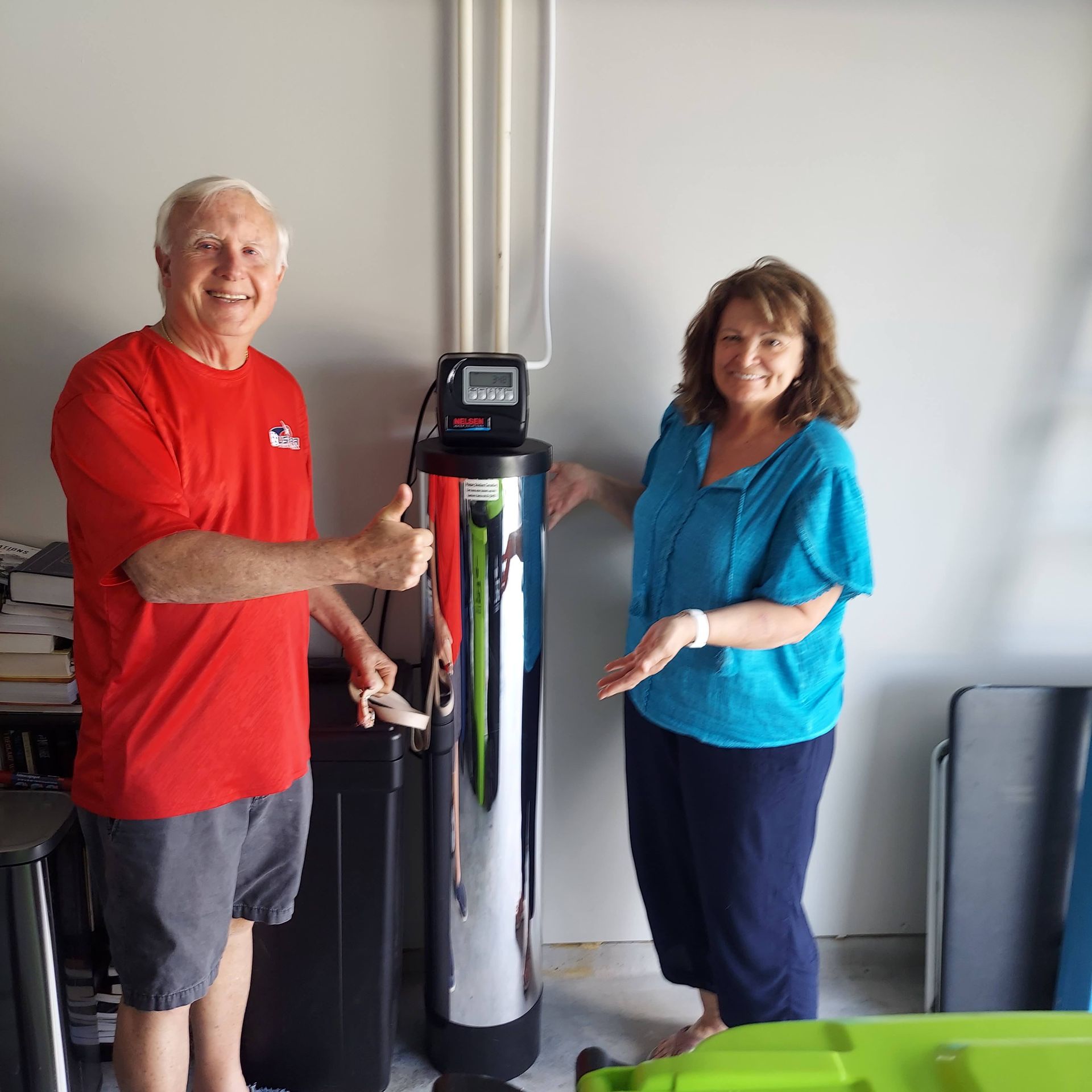 Man gives thumbs up, woman points, beside water softener in a garage.