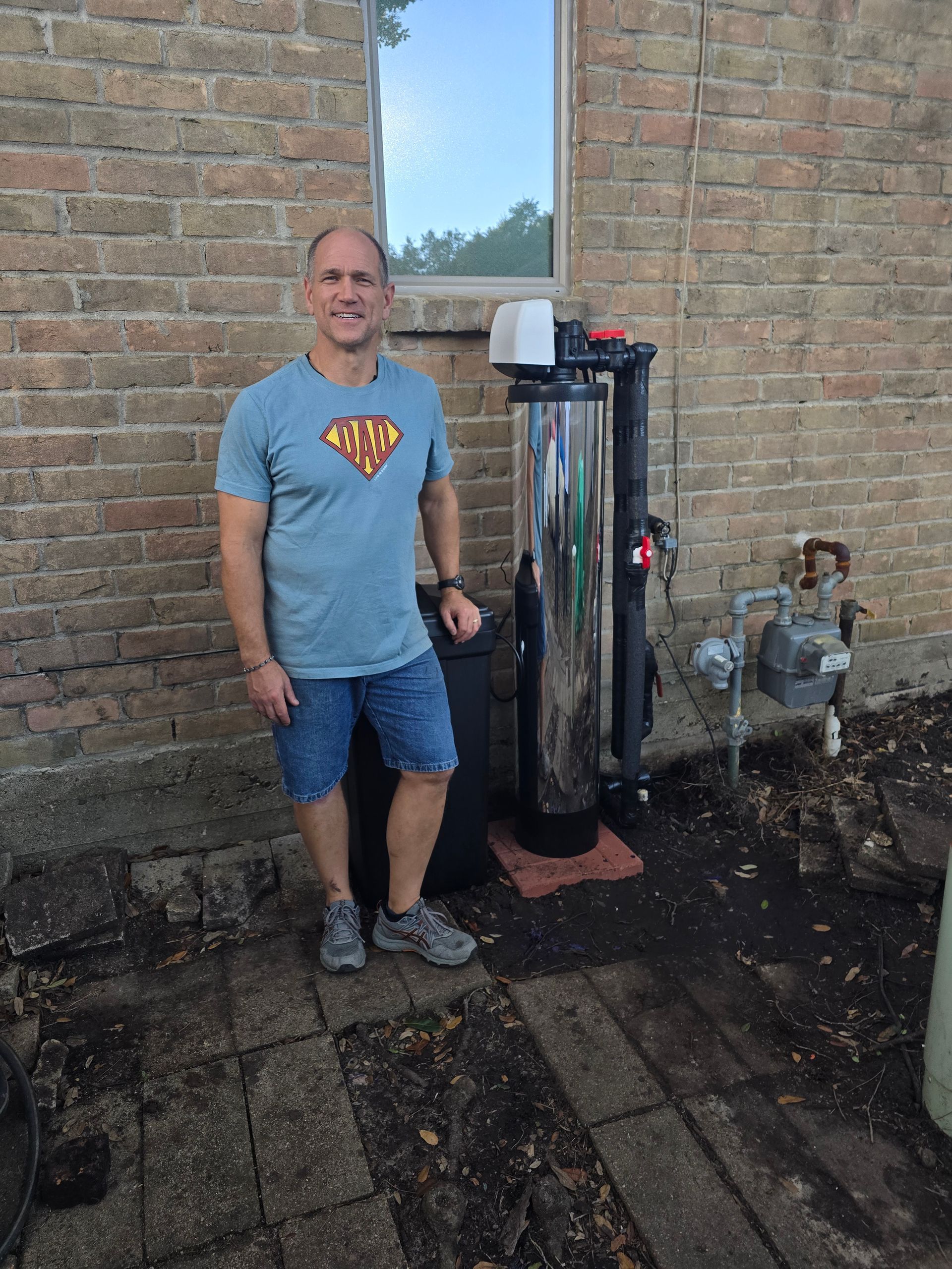Man smiles next to a water filtration system against a brick building.