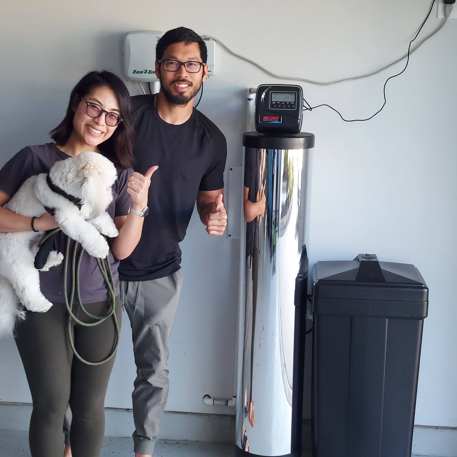 Couple with puppy thumbs up next to new water filtration system in a garage.