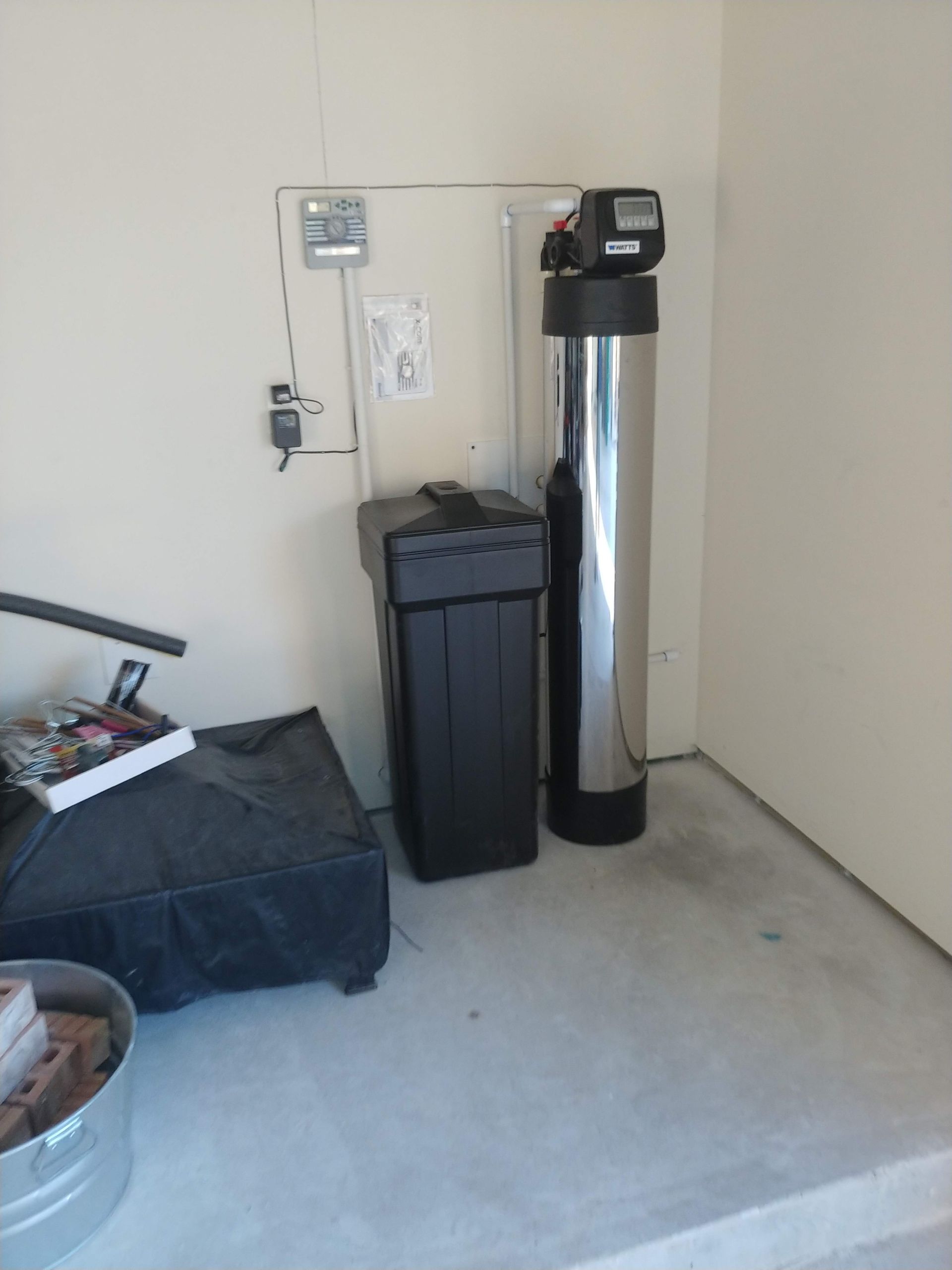 Water softener system in a garage setting with black tank, and tall chrome cylinder.