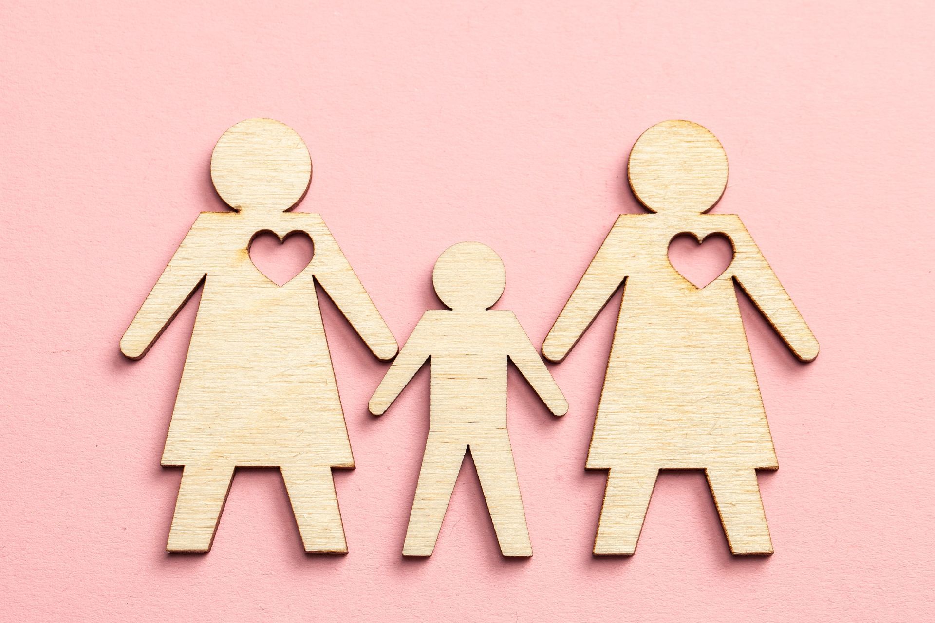 Three wooden figures holding hands on a pink background, with heart cutouts on the two larger figures