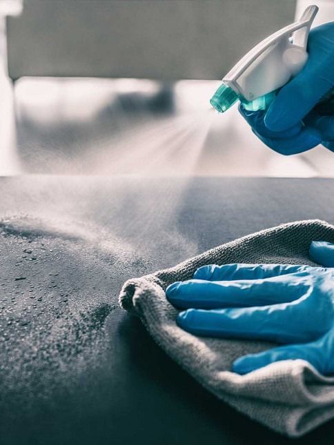 Hands in blue gloves spraying and wiping a dark surface with a cloth.