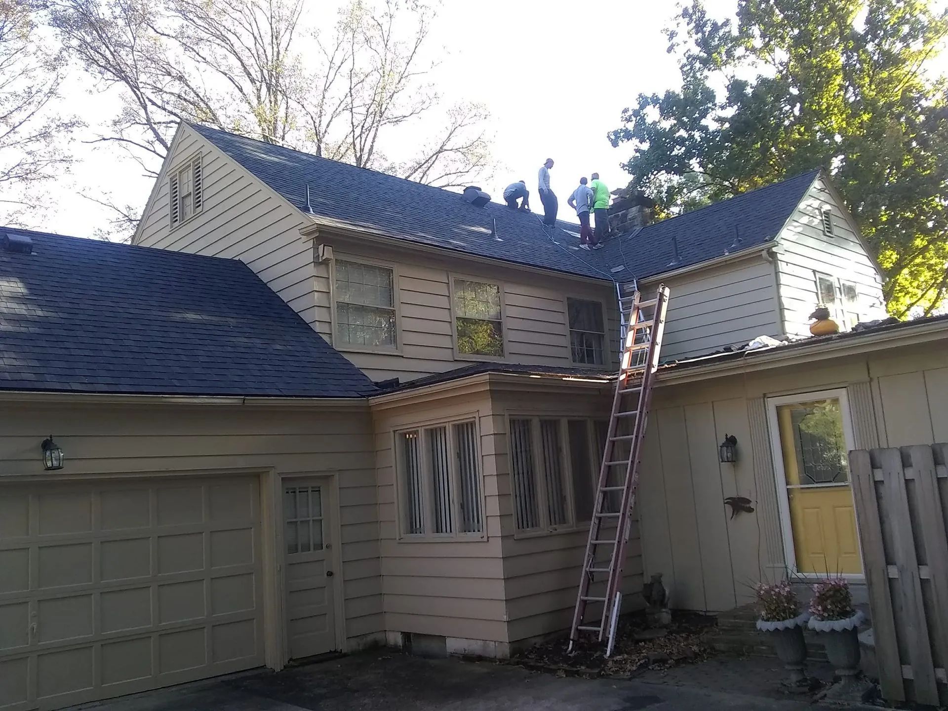 Workers on a roof with ladders outside a beige house and garage.