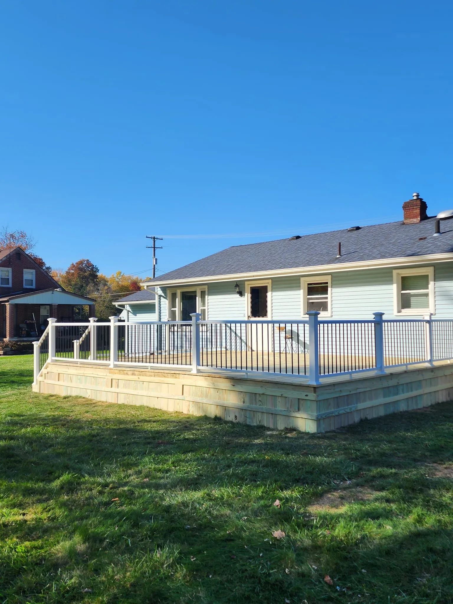 White house with a wooden deck and railing in a sunny grassy yard under a blue sky