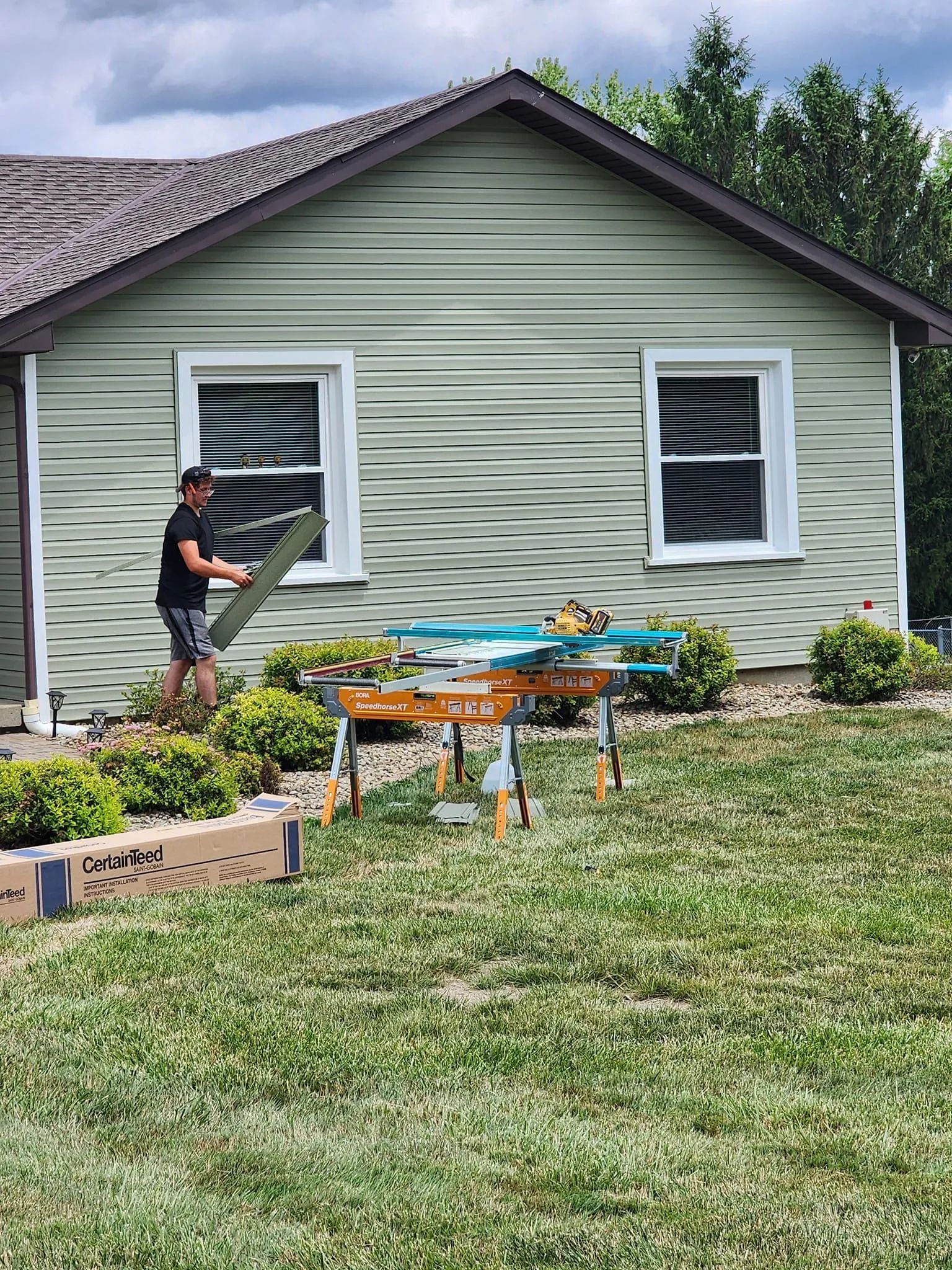 Man cutting siding on a table saw outside a house with green siding, cloudy sky.