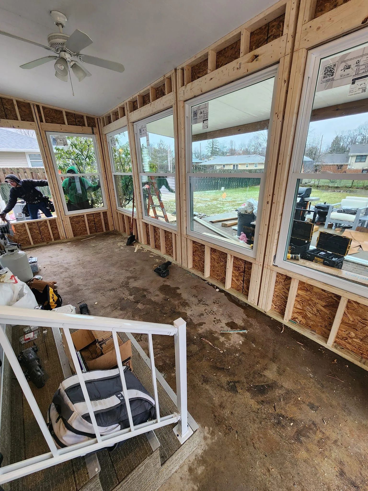 Construction workers install new windows in an open-walled residential sunroom with wooden framing and a ceiling fan.
