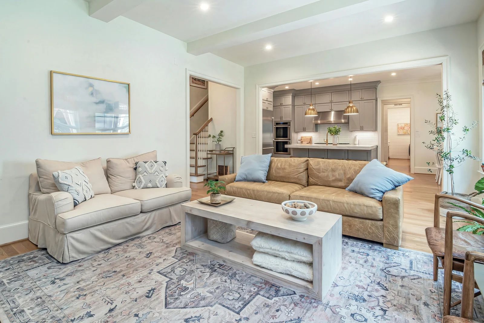 Bright living room with beige sofas, white coffee table, patterned rug, and open kitchen in the background