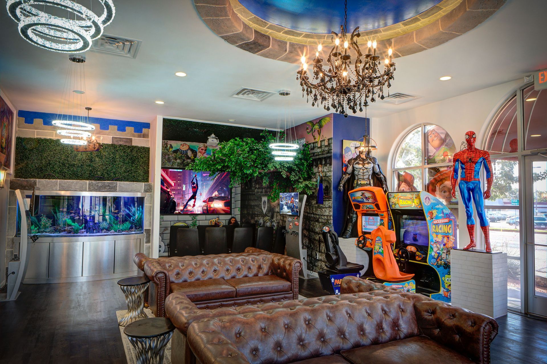 Upscale lounge with tufted sofas, chandelier, aquarium, greenery, and superhero mannequins on display.