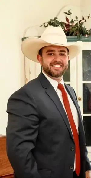 Smiling man in a white cowboy hat, dark suit, and red tie standing indoors.