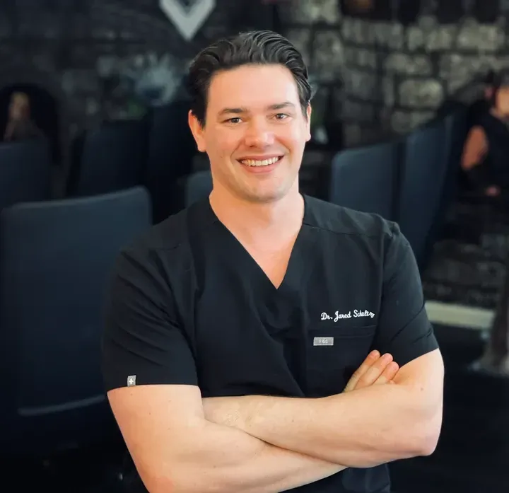 Smiling person in black scrubs with arms crossed, standing indoors against a dark, blurred background