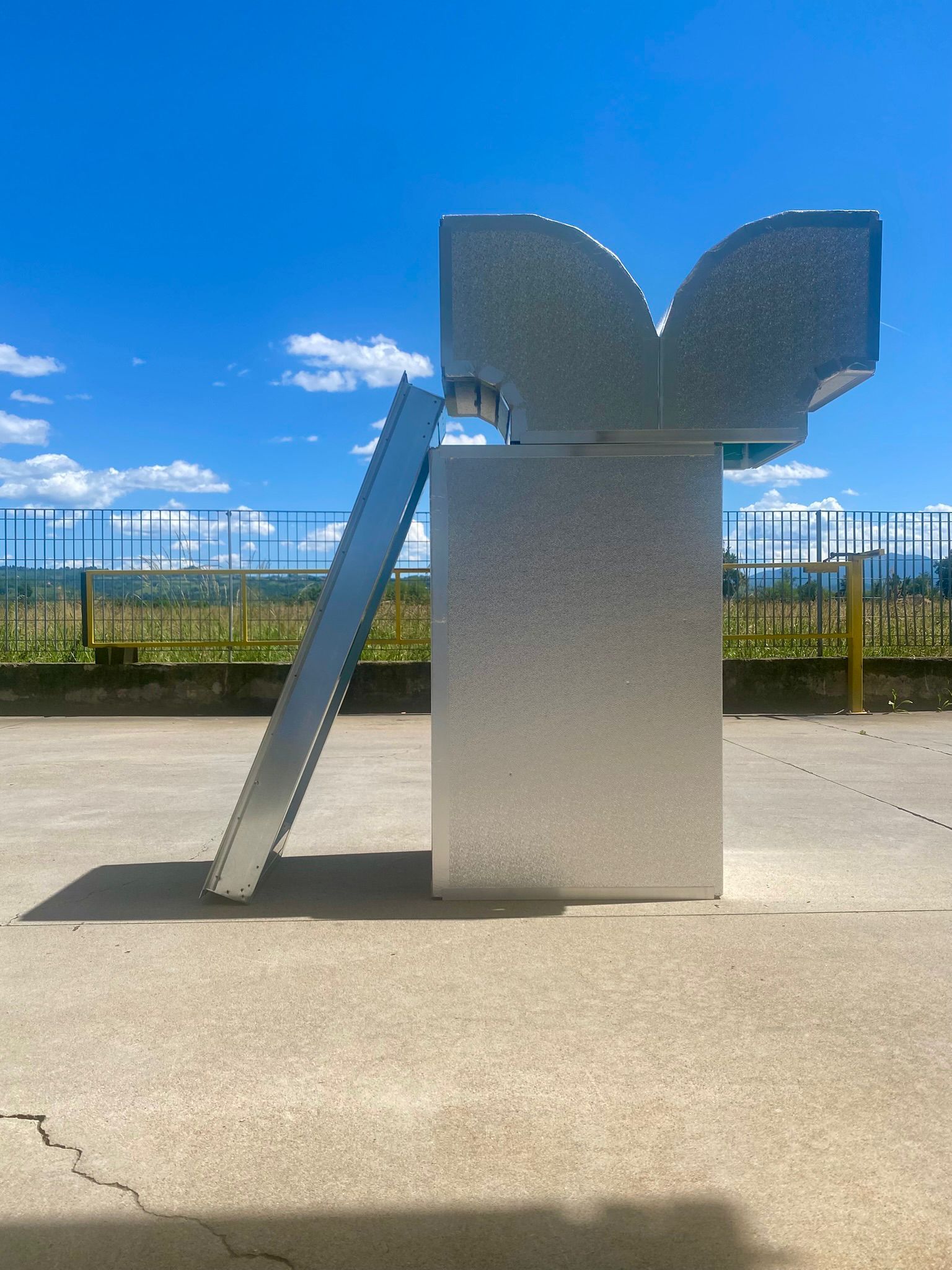 Scultura astratta moderna all'aperto, accanto a una recinzione, sotto un cielo azzurro brillante.