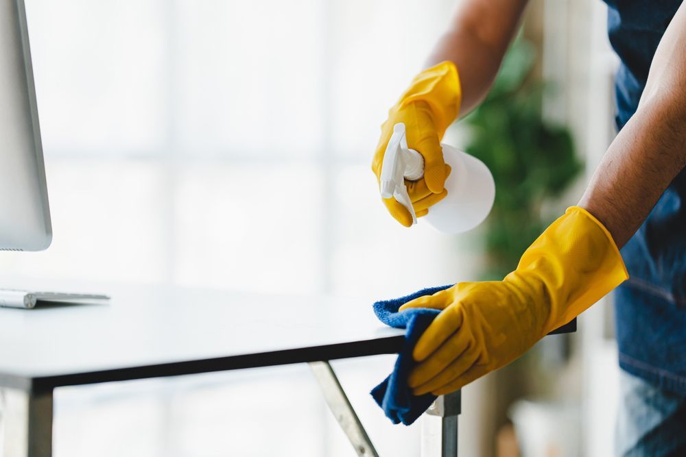 A Person Wearing Yellow Gloves Is Cleaning a Desk with A Cloth and Spray Bottle — Haydyn's Cleaning In Pialba, QLD