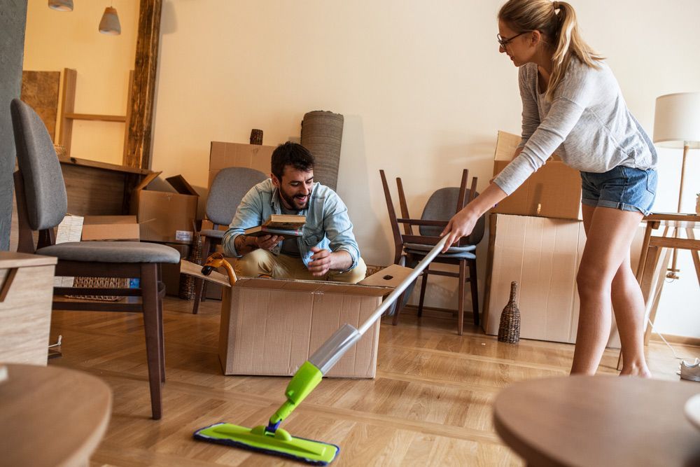 A Woman Is Mopping the Floor in A Living Room While a Man Sits in A Cardboard Box — Haydyn's Cleaning In Eli Waters, QLD