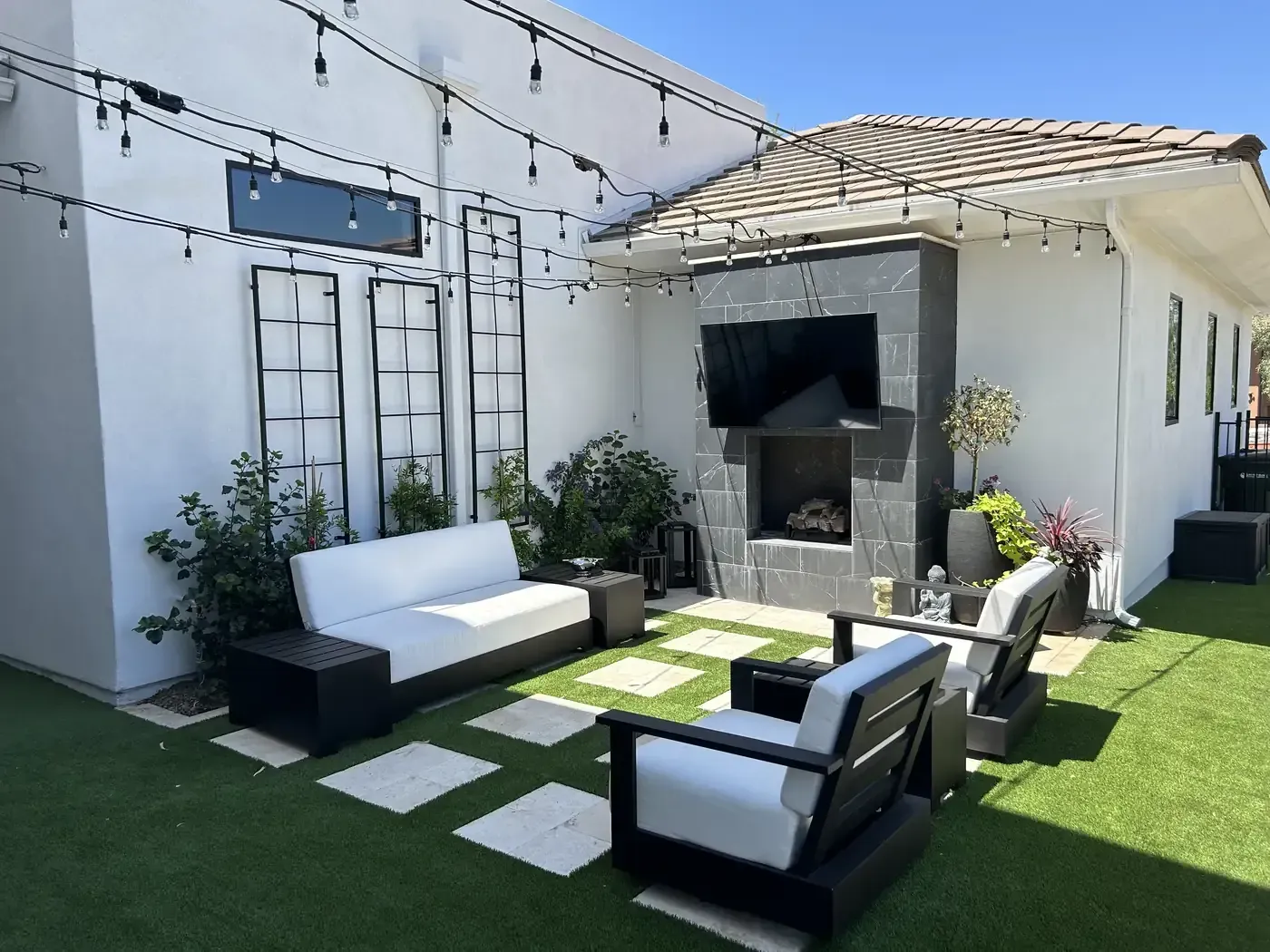 Modern patio with black and white outdoor seating, string lights, potted plants, and a wall-mounted TV
