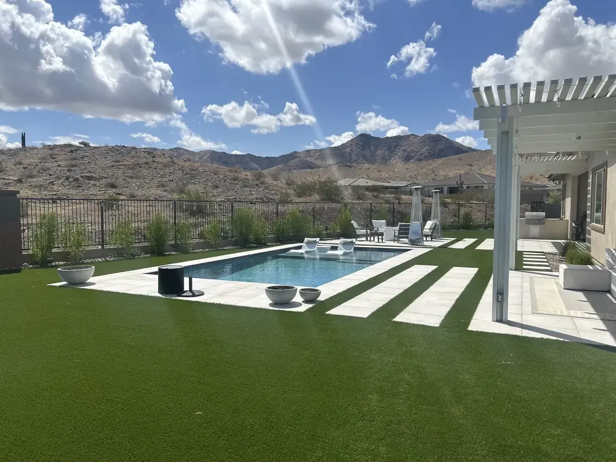 Modern backyard patio with pool, green lawn, white pergola, and mountain view under a cloudy sky