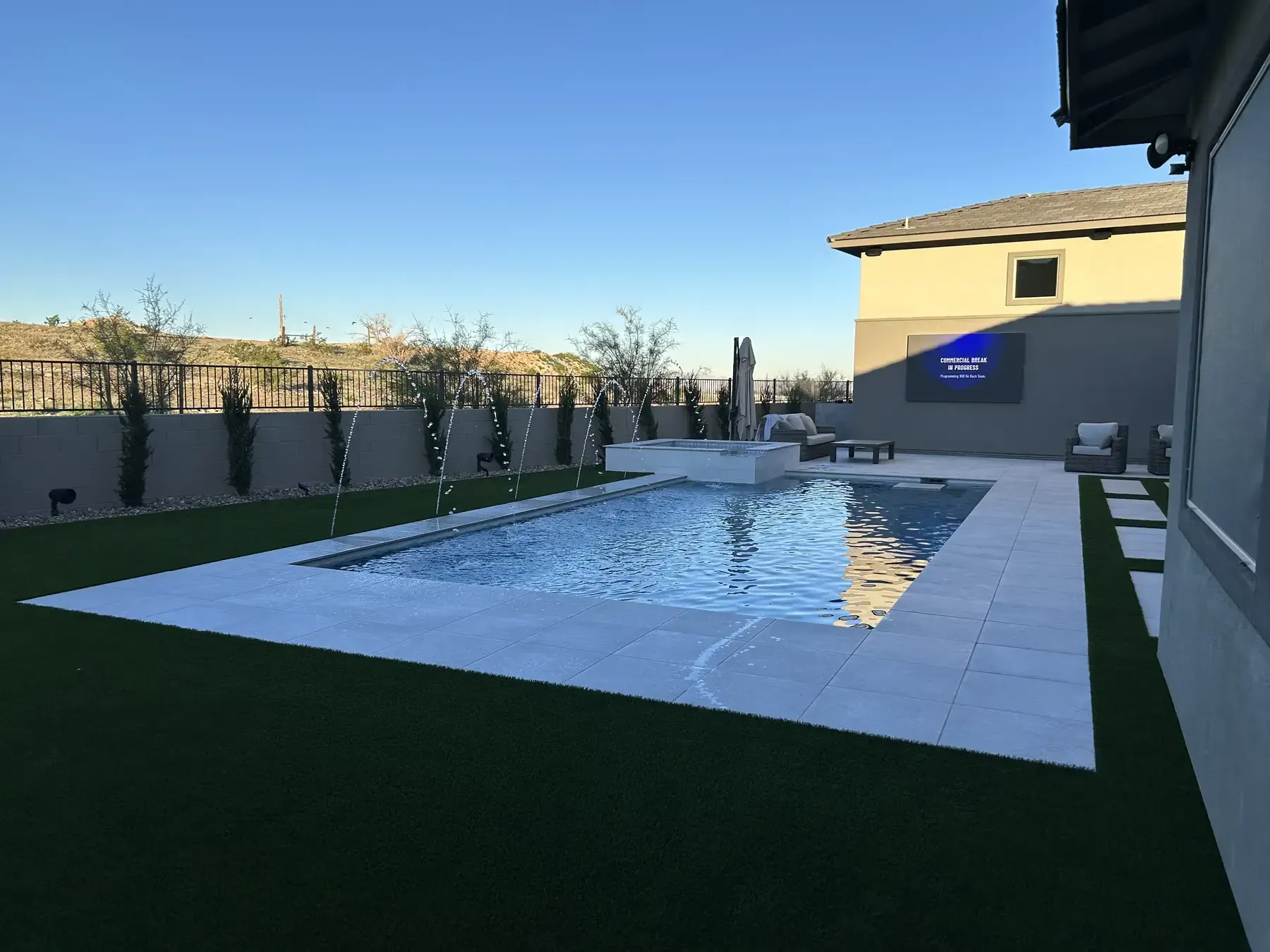 Modern backyard with a rectangular pool, white patio, and artificial grass beside a house at dusk