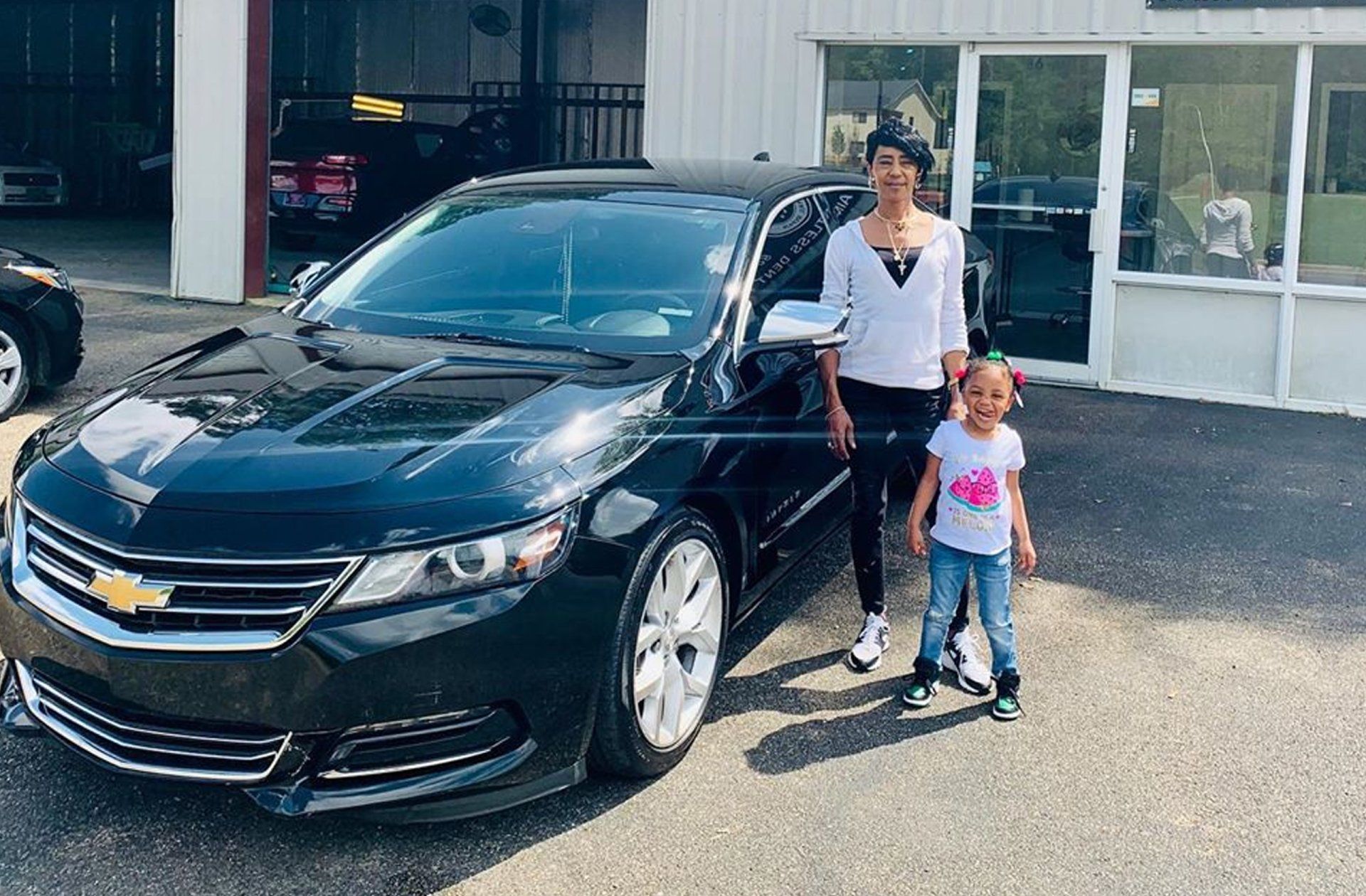 A woman and a little girl are standing in front of a black car.