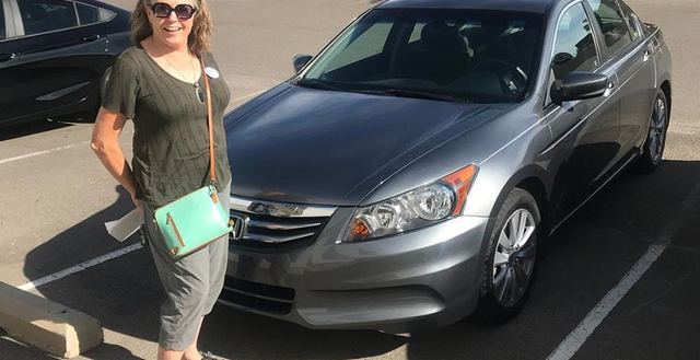 A woman is standing in front of a gray car in a parking lot.