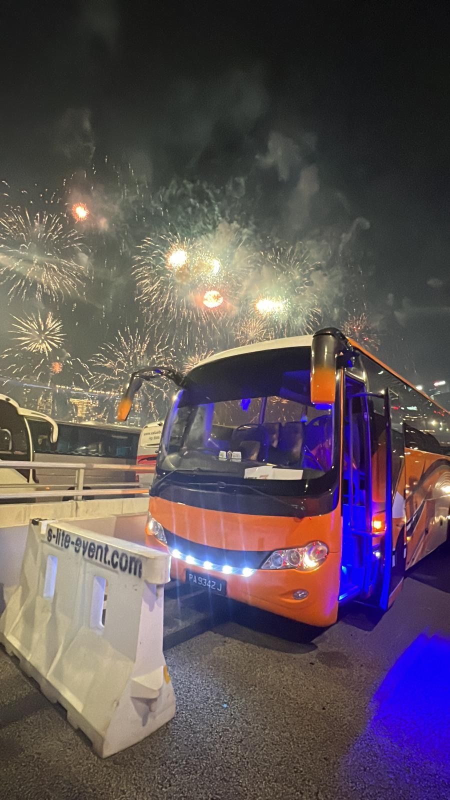 Orange bus parked with fireworks exploding in the night sky behind it.