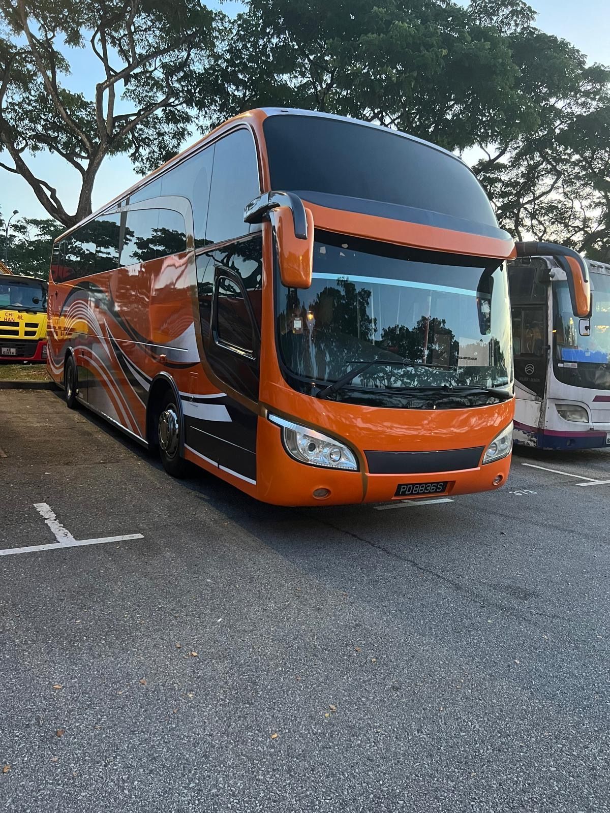 Orange coach bus parked in a lot beside other buses and trees