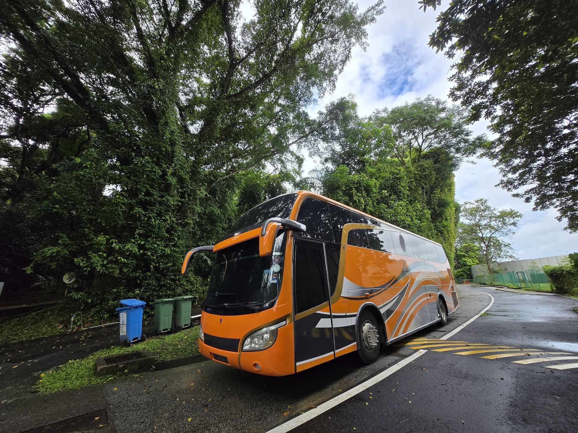 Orange and black tour bus parked on a wet road near green trees.