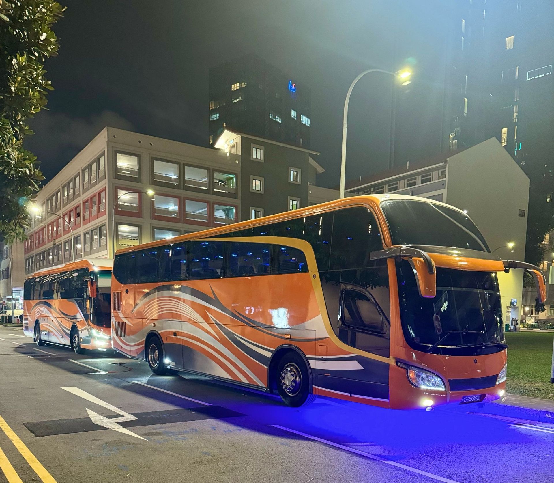 Two orange and black coaches parked at night on a city street with purple underglow lights