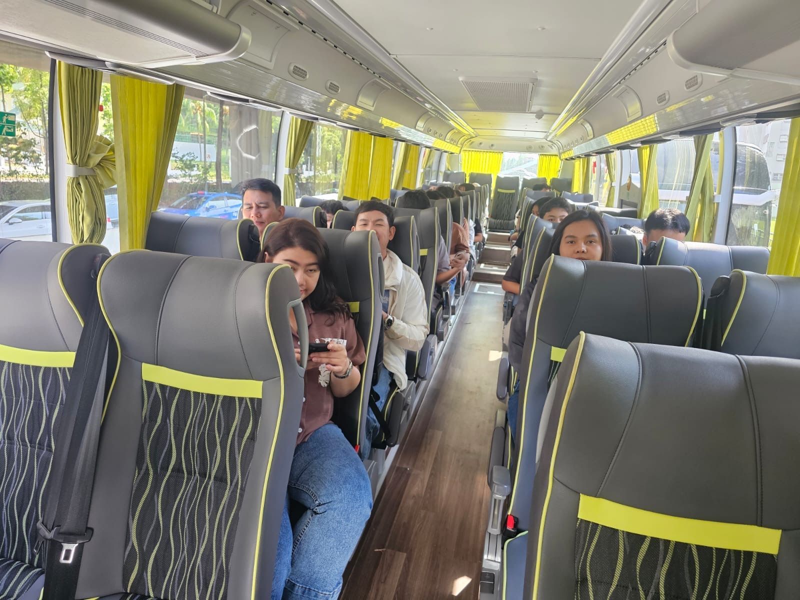 Passengers seated inside a modern bus with gray and yellow seats, looking at phones and out the windows.