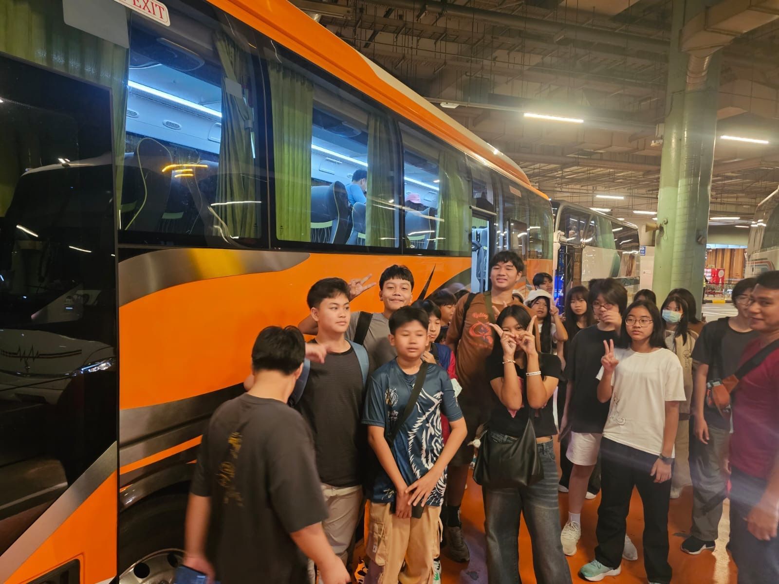 Group of students standing next to an orange and silver bus inside a garage, some smiling.