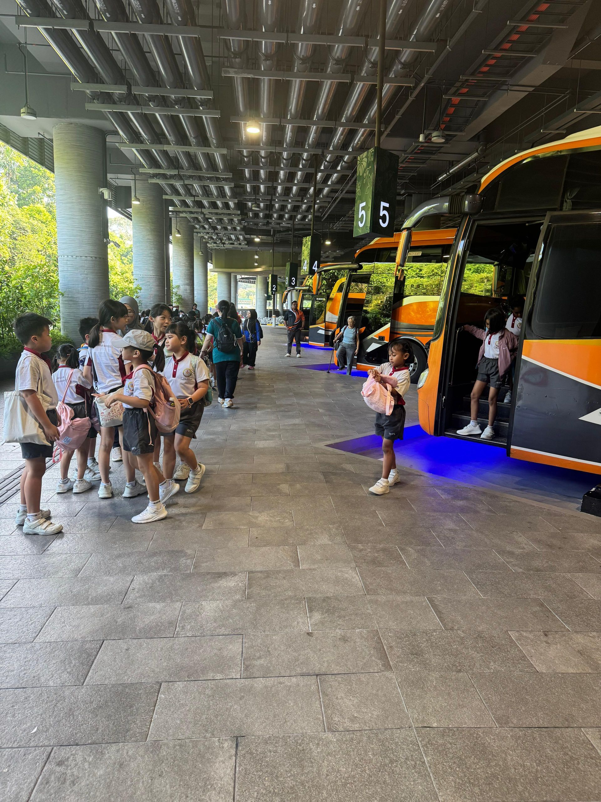 A group of children in white school uniforms stand by a line of yellow and black buses at a paved outdoor bus terminal.