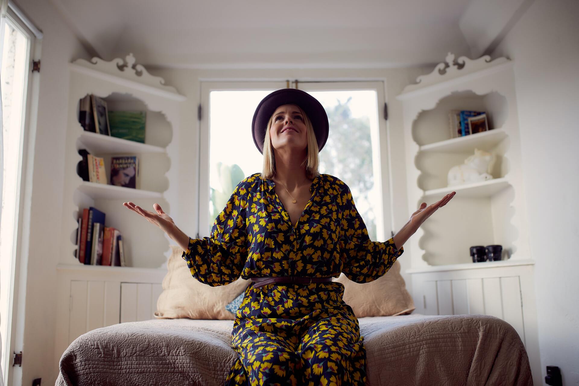 A woman in a floral dress and hat is sitting on a bed.