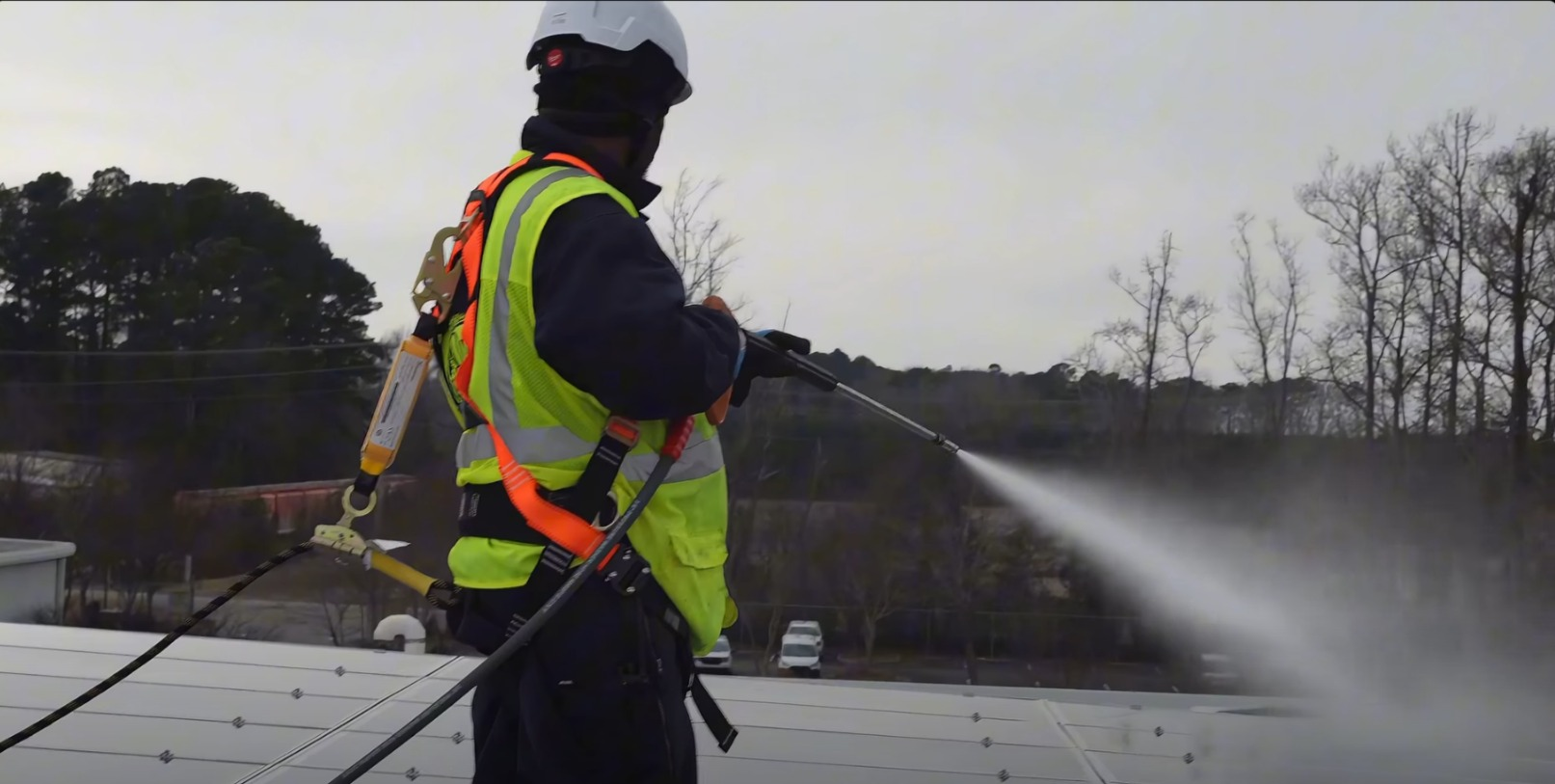 Worker in neon safety gear spraying water on a rooftop, with trees and overcast sky in the background.