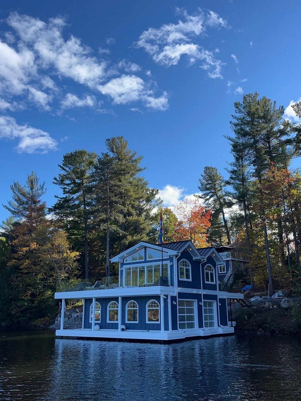 Blue lakeside cottage with white trim and porch, surrounded by pine trees under a bright sky.