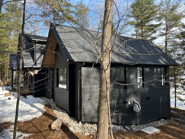 Black cabin with steep roof in snowy woods, viewed from the side among leafless trees.