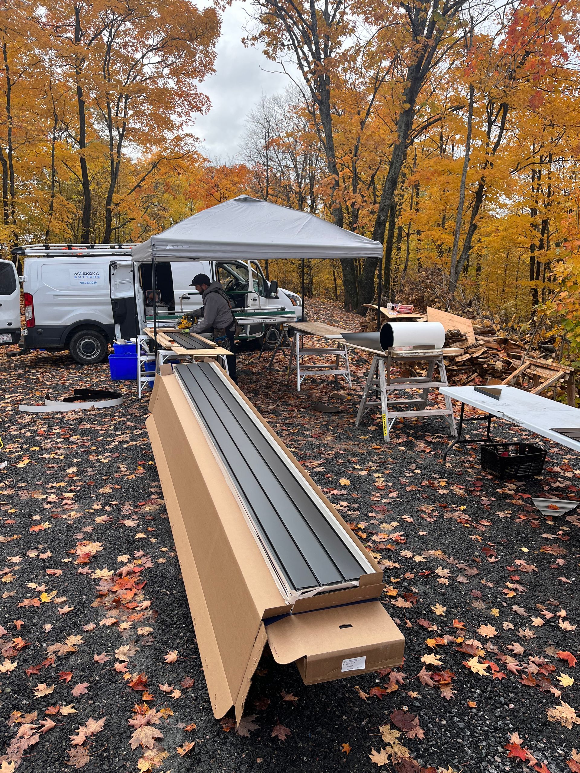 Outdoor worksite with canopy, truck, and long metal rails on cardboard in a wooded fall setting