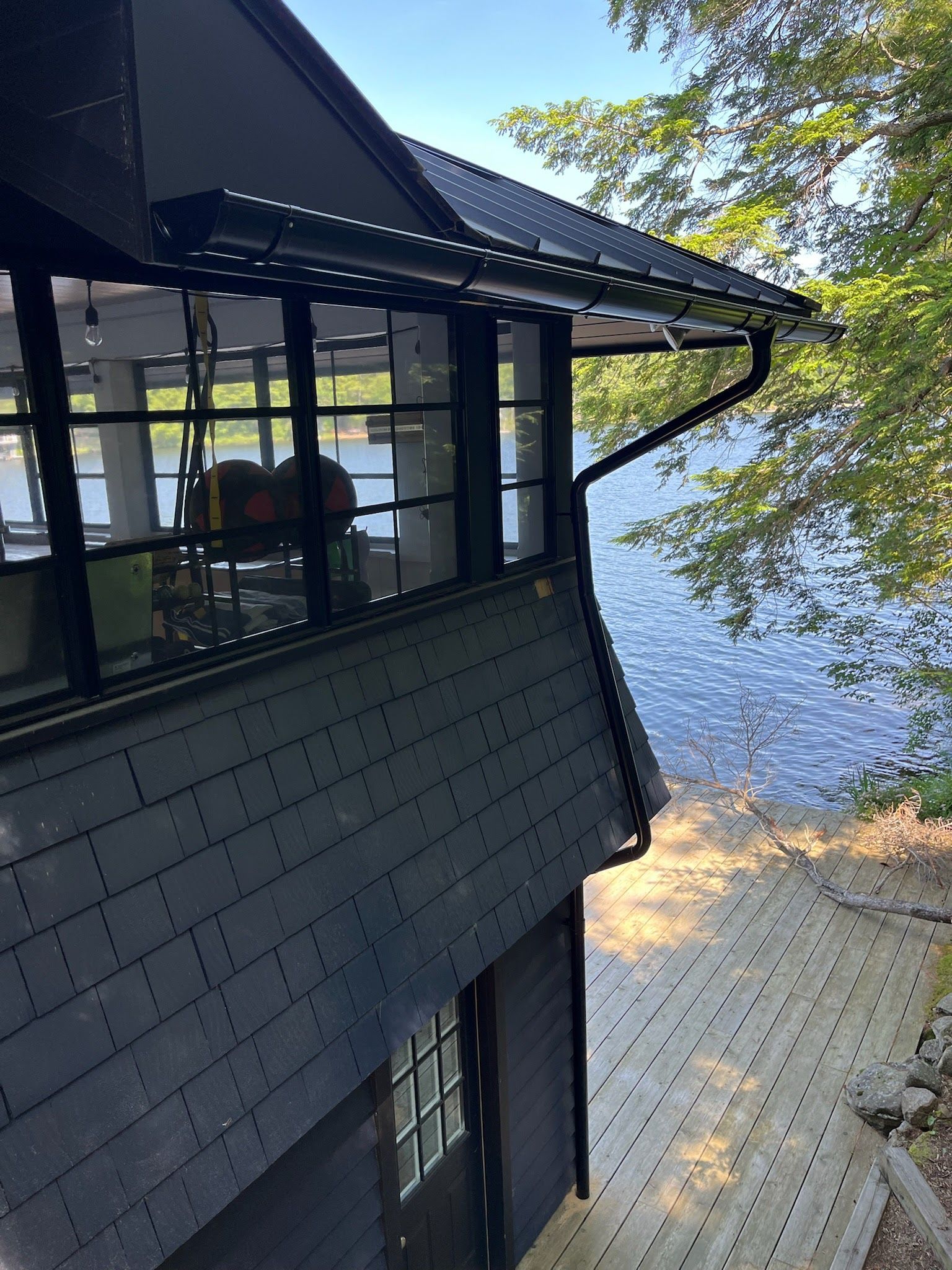 Exterior of a dark house beside a lake, with a paved patio and trees in sunlight