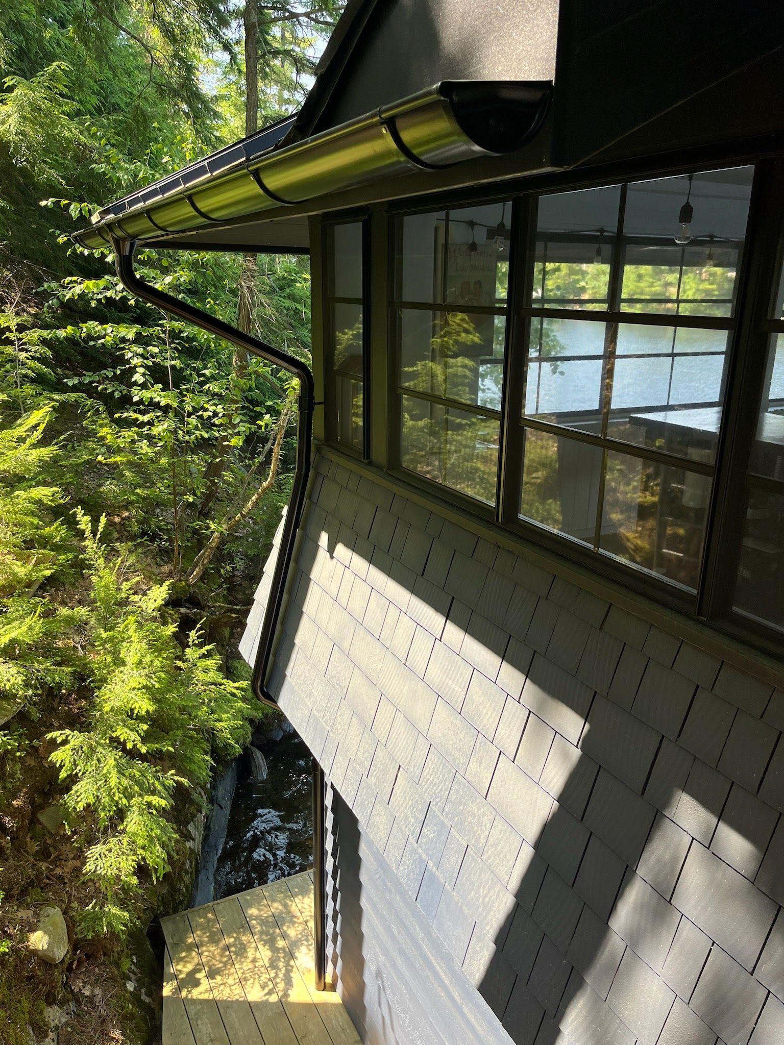Exterior of a house with large windows, roof eaves, and sunlight over a leafy hillside view