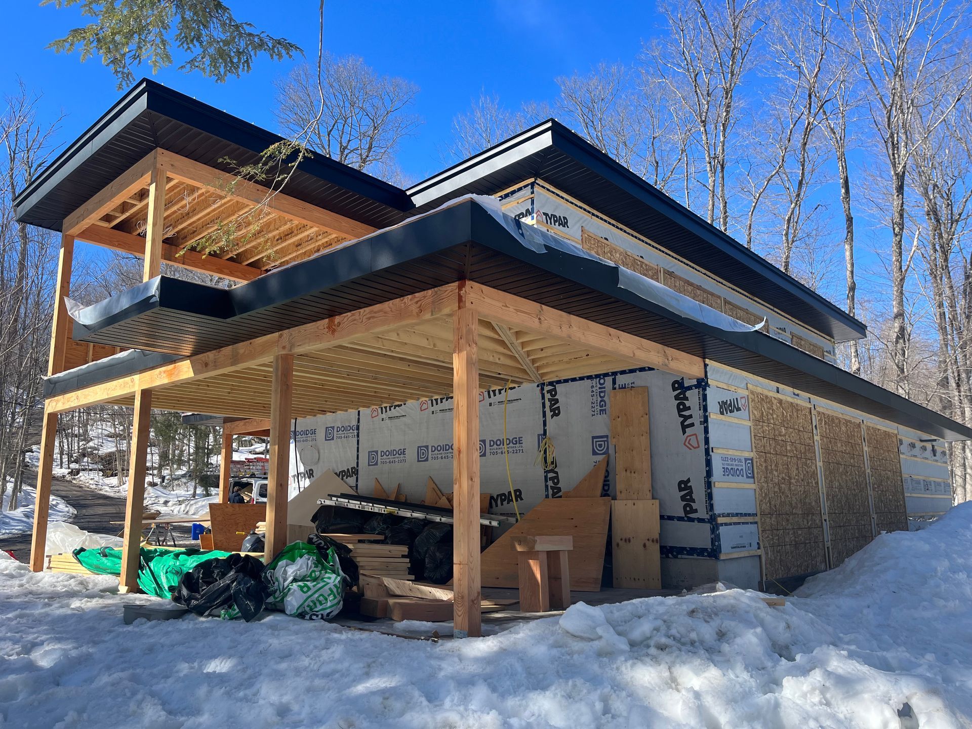 Modern house under construction in snowy woods, with exposed framing and black roof under a blue sky