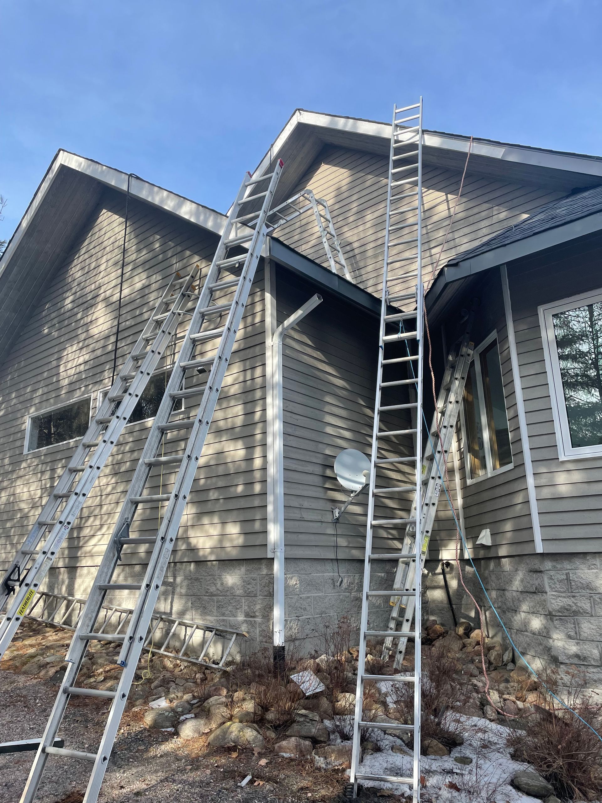 Three ladders leaning against a house exterior with stone and siding, likely for roof or gutter work