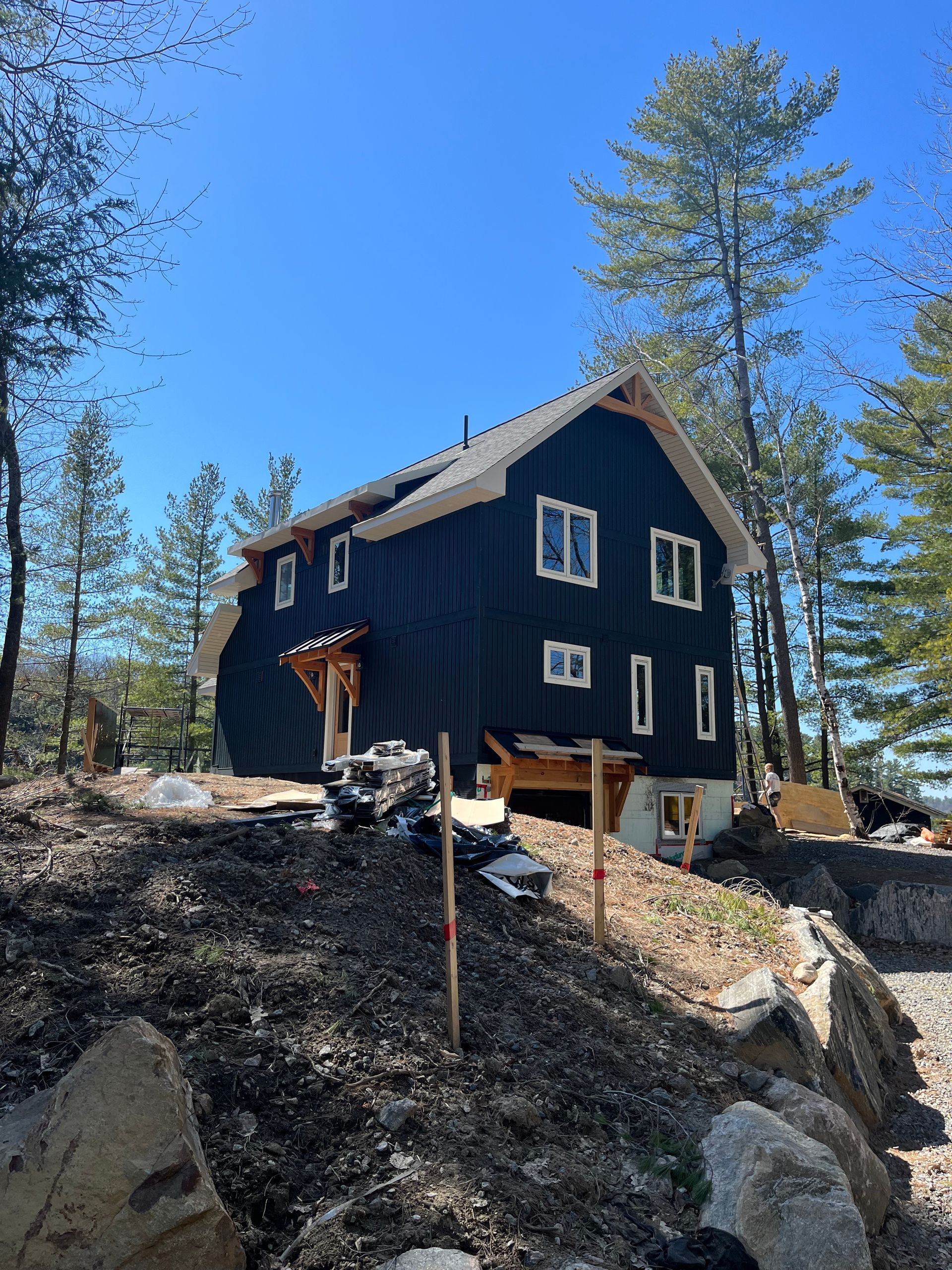 Dark blue cabin on a rocky hillside surrounded by pine trees under a clear blue sky
