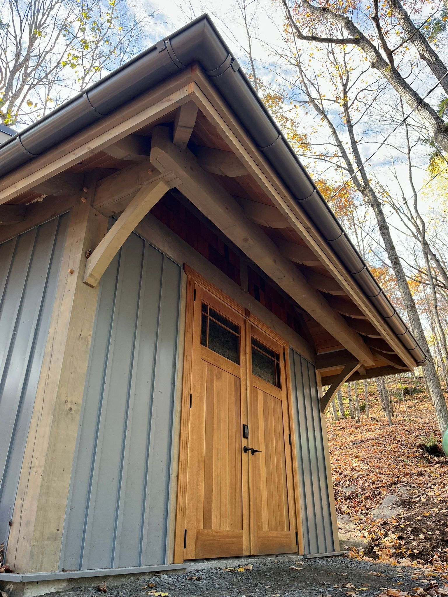 Small rustic shed with blue siding and wooden double doors beneath a steep roof in a wooded area.