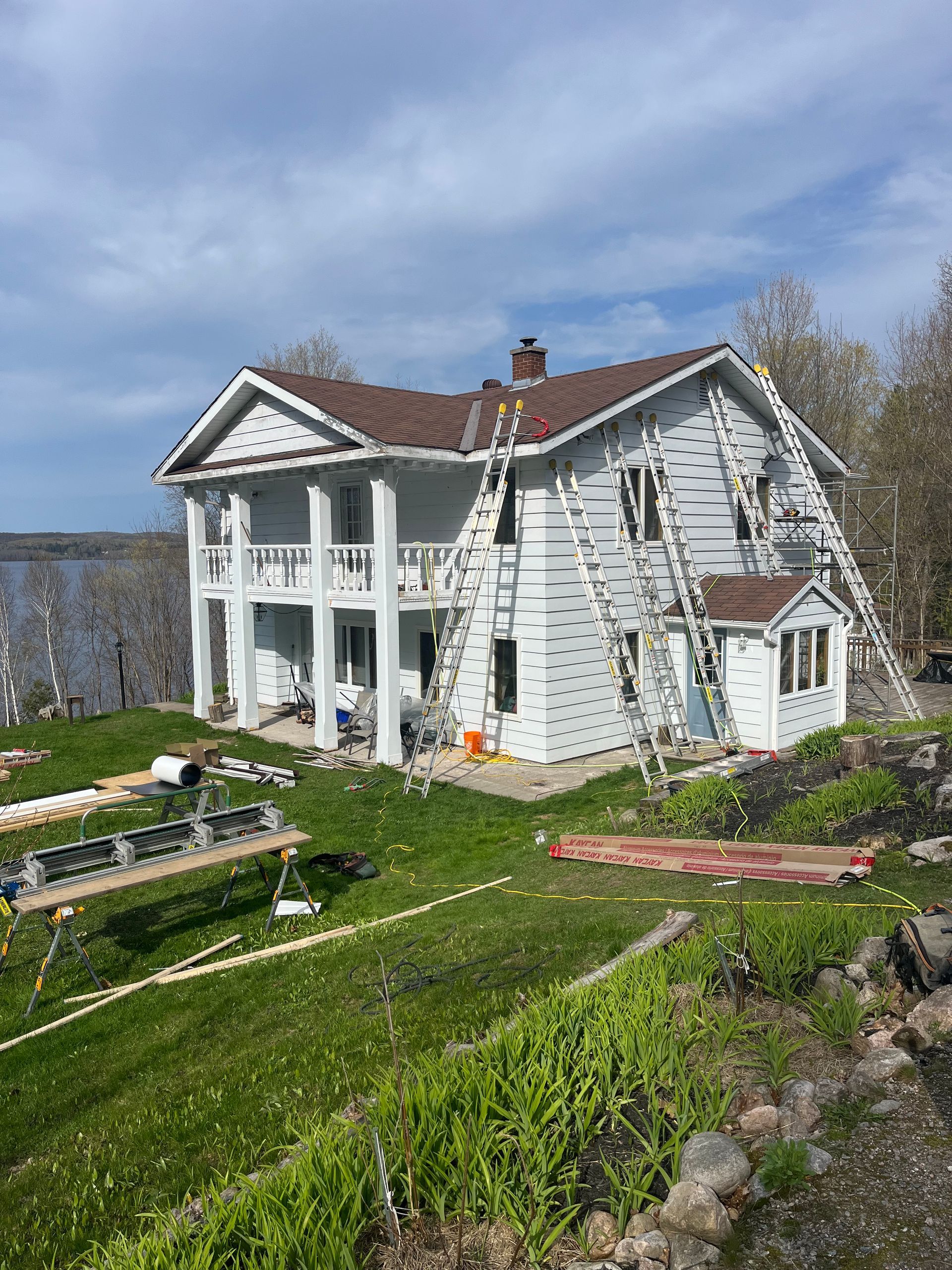 White two-story house with red roof by the water, with a ladder leaning against it and a garden in front