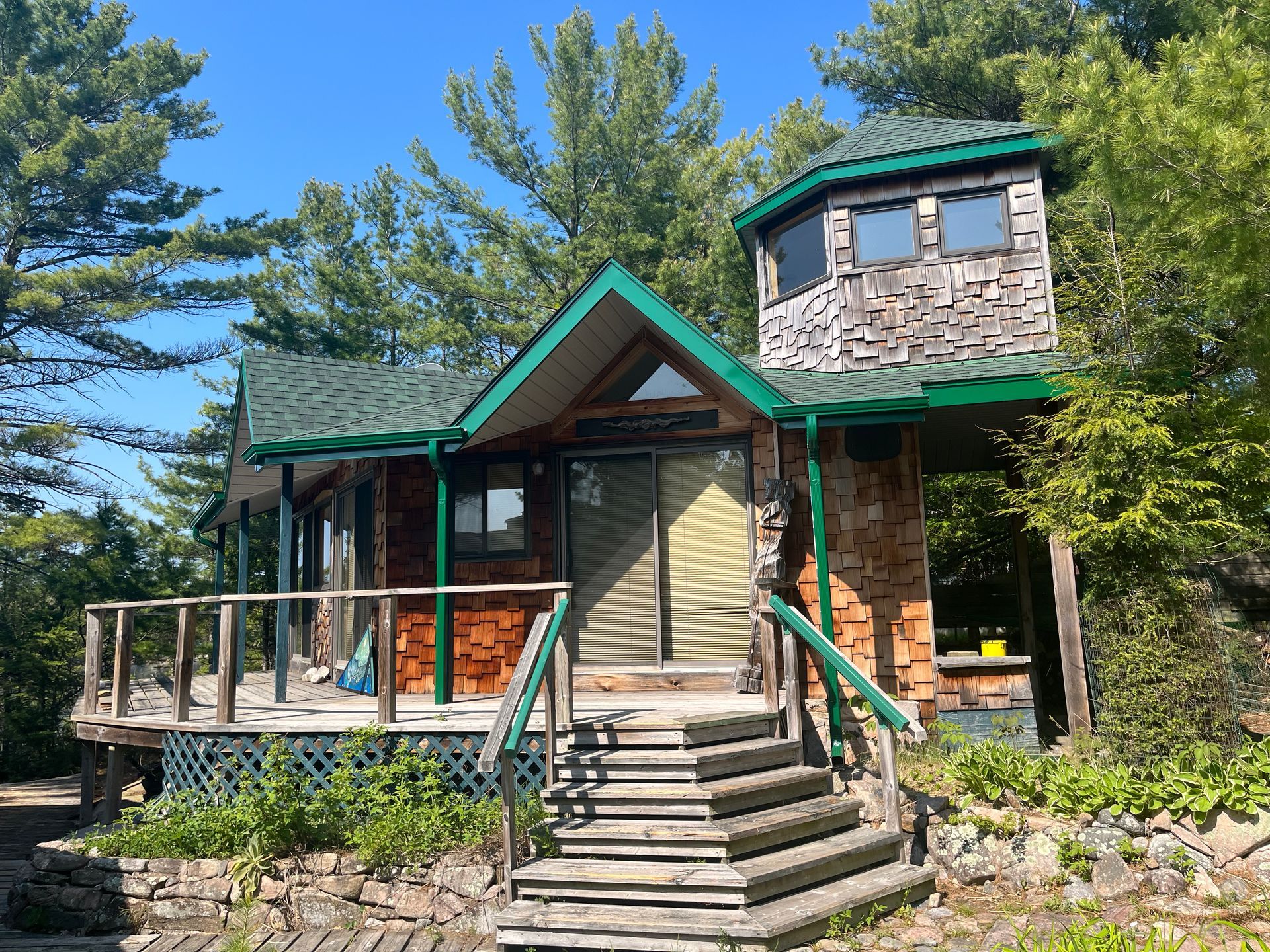 Stone cabin with green roof and porch, surrounded by trees under a clear blue sky