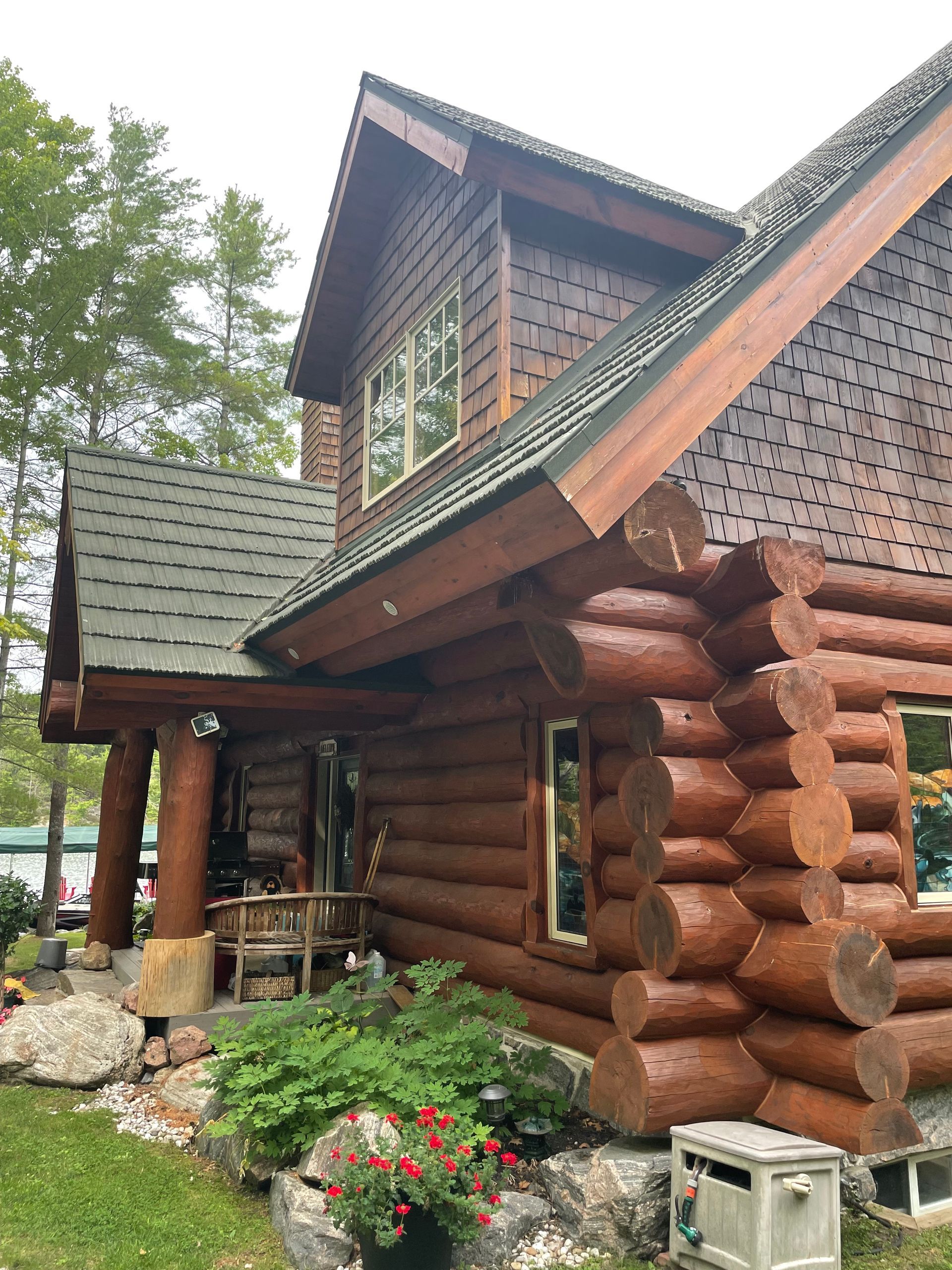 Rustic log cabin with a green shingled roof, flower bed, and a small porch in a wooded setting
