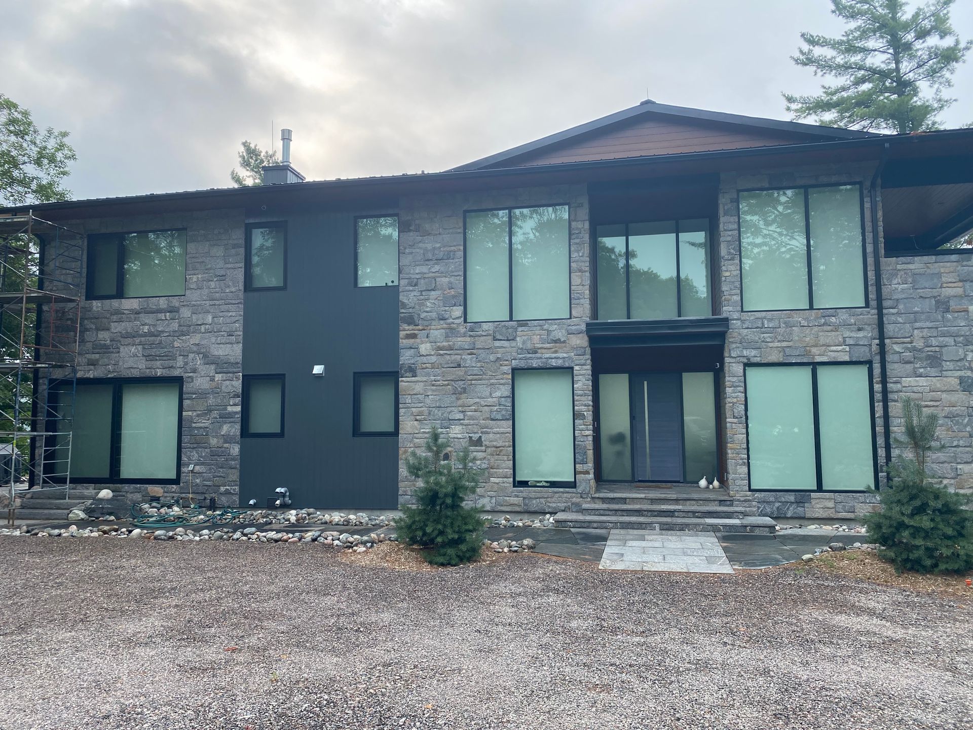 Modern two-story house with dark siding, stone center, large windows, and gravel driveway under cloudy sky