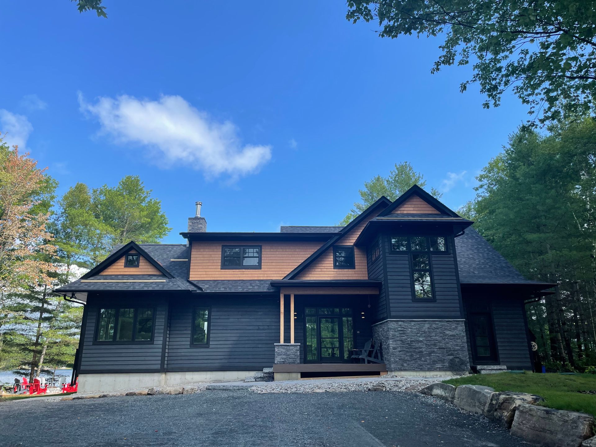 Dark wooden house with stone foundation and large windows, surrounded by trees under a blue sky
