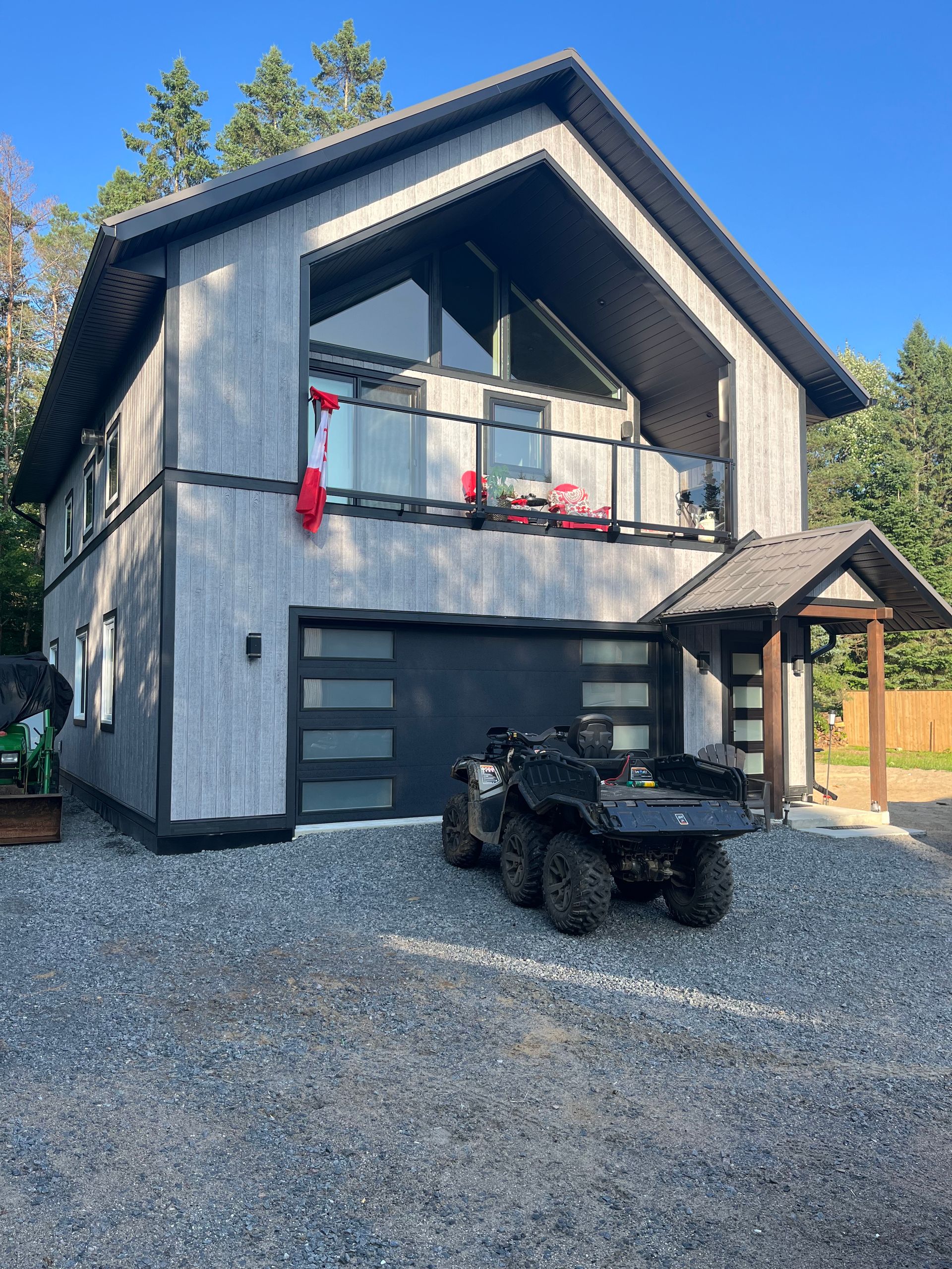 Modern two-story gray house with balcony, garage, and black ATV parked on gravel driveway