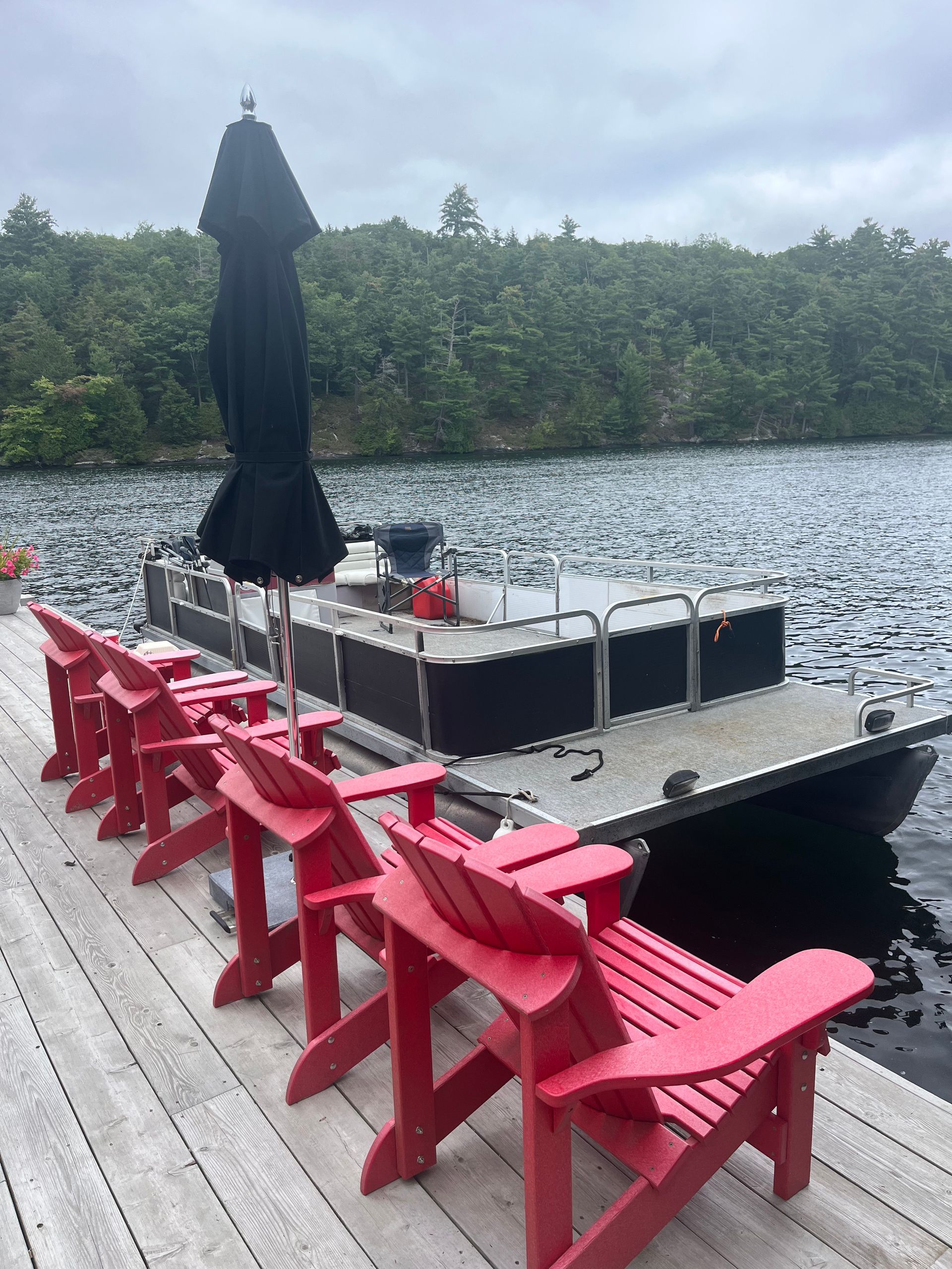 Red Adirondack chairs and a black umbrella on a lakeside dock with forested shoreline.