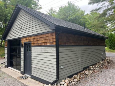 Small gray detached garage with black trim and shingled roof on a gravel driveway, surrounded by trees.