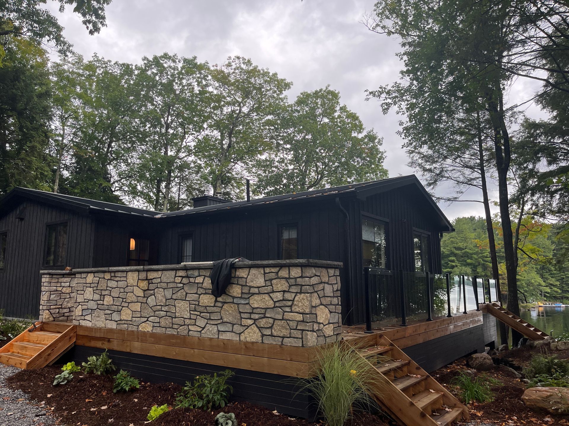 Modern black cabin with stone foundation on a wooded lakeside lot, viewed from a deck.